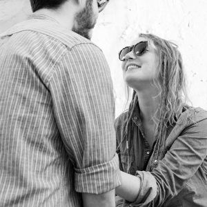 En una fotografía en blanco y negro, una mujer con gafas de sol sonríe a un hombre con una camisa de rayas, encarnando el espíritu de "Un paseo de fotografía en pareja en Granada". - ForTwo - Planes para parejas