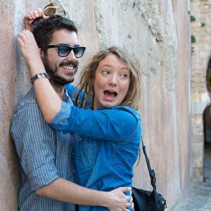 Una mujer sorprendida y un hombre sonriente se abrazan alegremente contra un muro de piedra, capturando un momento alegre mientras disfrutan de "Un paseo de fotografía en pareja en Granada. - ForTwo - Planes para parejas