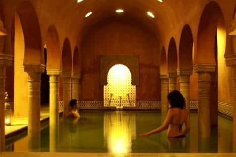 Los huéspedes del MIMMA - Baño árabe con masaje en Hammam Al Ándalus Granada se relajan en una piscina cubierta arqueada, cálidamente iluminada y rodeada de columnas y diseños intrincados, saboreando el ambiente sereno de un baño árabe tradicional. - ForTwo - Planes para parejas
