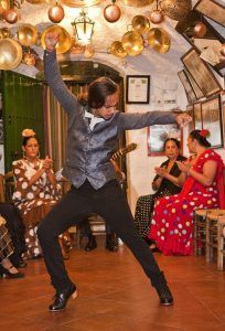 En una sala con poca luz, una bailarina de flamenco actúa apasionadamente durante un Espectáculo de flamenco en la Zambra María la Canastera, mientras mujeres sentadas con vestidos de lunares aplauden. - ForTwo - Planes para parejas
