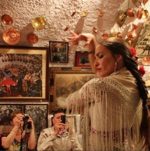 En una sala bellamente adornada, una bailaora de flamenco realiza con pasión un espectáculo impresionante mientras dos espectadores cautivados capturan el momento, que recuerda al vibrante "Espectáculo de flamenco en la Zambra María la Canastera". - ForTwo - Planes para parejas