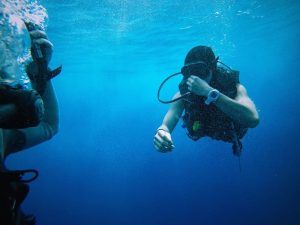 Dos buceadores bajo el agua, participando en la experiencia Oferta: Bautismo de buceo (Granada), ajustan su equipo en medio de las cristalinas aguas azules. - ForTwo - Planes para parejas