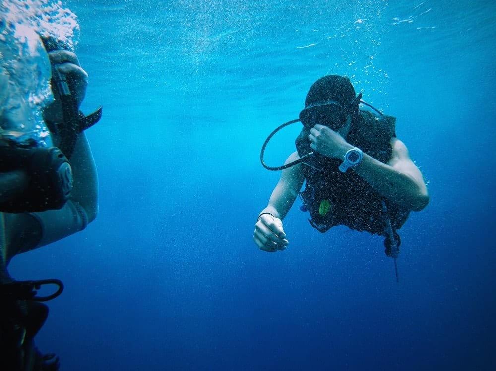 Dos buceadores bajo el agua, participando en la experiencia Oferta: Bautismo de buceo (Granada), ajustan su equipo en medio de las cristalinas aguas azules. - ForTwo - Planes para parejas