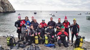 Buceadores con sombreros festivos posan en una pintoresca playa de guijarros con el equipo de buceo listo y el sereno mar de Granada como telón de fondo, celebrando una experiencia de Oferta: Bautismo de buceo (Granada). - ForTwo - Planes para parejas