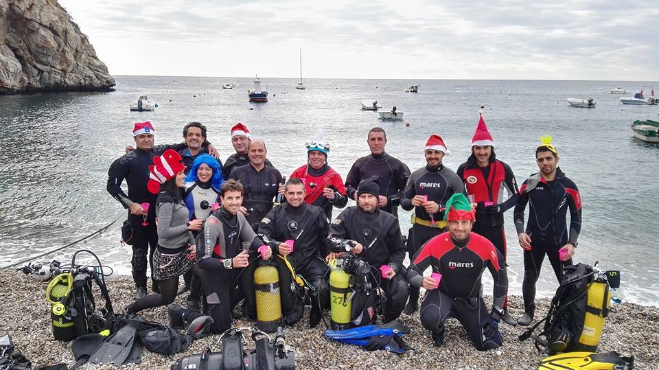 Buceadores con sombreros festivos posan en una pintoresca playa de guijarros con el equipo de buceo listo y el sereno mar de Granada como telón de fondo, celebrando una experiencia de Oferta: Bautismo de buceo (Granada). - ForTwo - Planes para parejas
