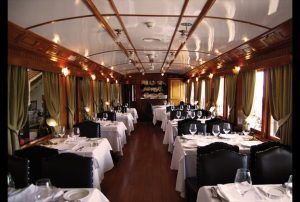 Elegante vagón comedor que recuerda al Restaurante El Vagón de Beni, con manteles blancos, mesas impecablemente puestas, cortinas verdes y cálidos paneles de madera. Ideal para un romántico viaje en tren a Madrid. - ForTwo - Planes para parejas