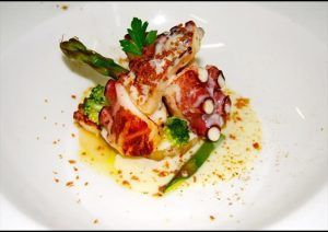Pulpo a la parrilla acompañado de espárragos, brócoli y salsa cremosa, coronado con perejil en un plato blanco en el Restaurante El Vagón de Beni - Un romántico tren en Madrid. - ForTwo - Planes para parejas