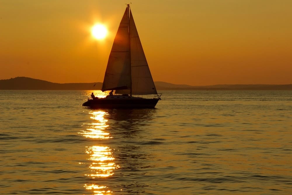 Un velero se recorta contra una puesta de sol, reflejándose en las aguas de Almuñécar, capturando la serena esencia de un tranquilo paseo romántico en barco en la Herradura y Almuñécar. - ForTwo - Planes para parejas