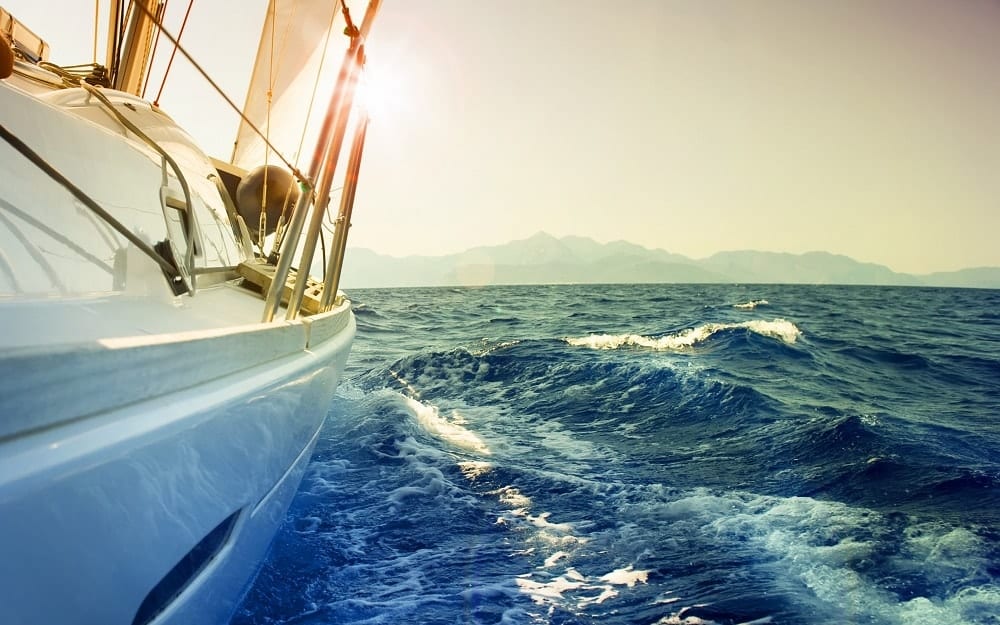 Un velero del "Romántico paseo en barco en la Herradura y Almuñécar" se desliza sobre el océano con la luz del sol reflejándose en el agua, montañas en la distancia y un cielo azul claro arriba. - ForTwo - Planes para parejas