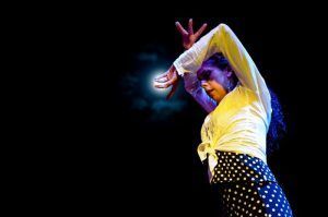 Una persona con un top blanco y una falda de lunares baila apasionadamente bajo un foco, capturando la esencia de "Espectáculo de flamenco en los Jardines de Zoraya" contra un fondo oscuro. - ForTwo - Planes para parejas