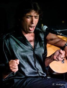 Una persona canta apasionadamente ante un micrófono, apretando los puños y vistiendo una camisa negra brillante que brilla con la energía vibrante que recuerda al Espectáculo de flamenco en los Jardines de Zoraya. - ForTwo - Planes para parejas