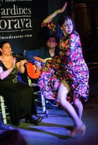 En el Espectáculo de flamenco en los Jardines de Zoraya, una apasionada bailarina con un vestido floral actúa mientras el guitarrista rasguea y el público aplaude, creando una experiencia inolvidable. - ForTwo - Planes para parejas