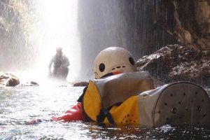 Una persona con traje de neopreno y casco flota tranquilamente en el Río Verde cerca de una impresionante cascada, representando la emoción del Barranquismo en Granada - Ruta completa Río Verde. Al fondo, otro aventurero disfruta de la encantadora belleza de esta joya escondida. - ForTwo - Planes para parejas