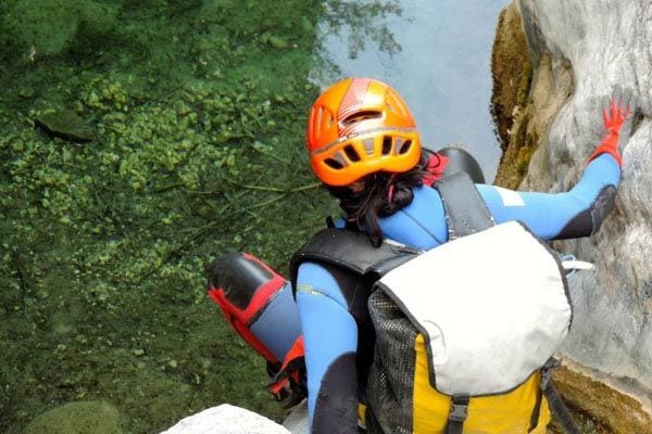 Vestido con un traje de neopreno, casco, mochila y guantes rojos, una persona sube con entusiasmo las rocas cerca de las aguas cristalinas de Río Verde en Granada para vivir una aventura con el Barranquismo en Granada - Ruta completa Río Verde. - ForTwo - Planes para parejas