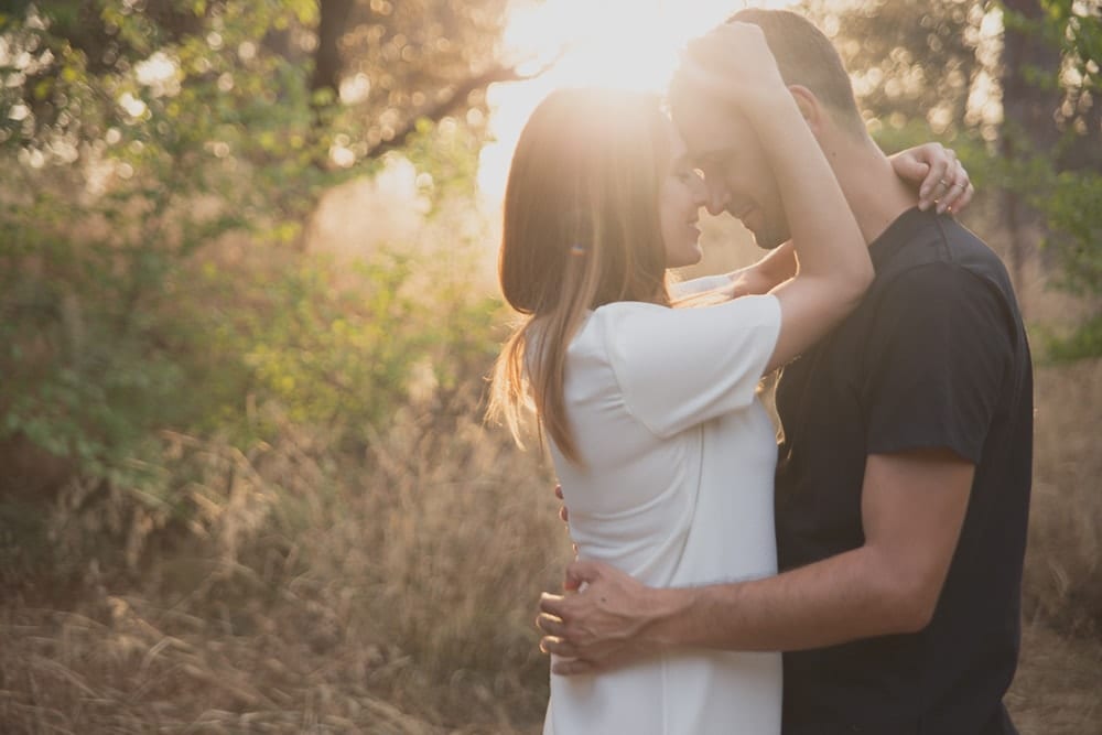 En el bosque iluminado por el sol, una pareja se abraza entre árboles y pasto con un atardecer de fondo, un escenario perfecto para una Sesión de fotografía en pareja de Carlos Fierro. - ForTwo - Planes para parejas