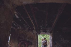 En una cautivadora "Sesión de fotografía en pareja por Carlos Fierro", una pareja se abraza en la entrada de un edificio oscuro y desgastado con vegetación visible afuera. - ForTwo - Planes para parejas