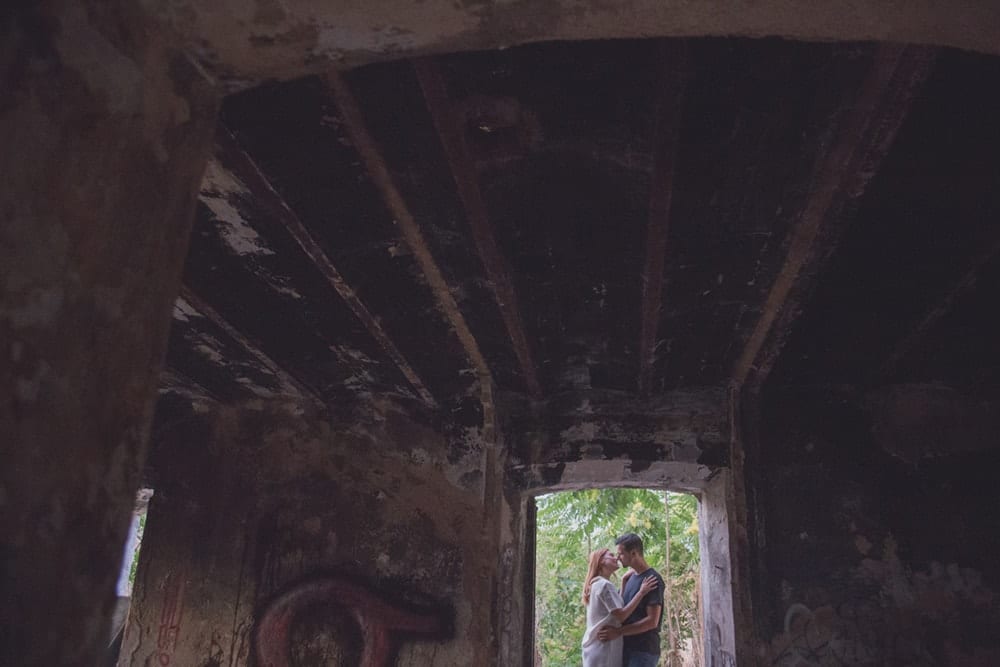En una cautivadora "Sesión de fotografía en pareja por Carlos Fierro", una pareja se abraza en la entrada de un edificio oscuro y desgastado con vegetación visible afuera. - ForTwo - Planes para parejas