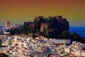 El castillo en la cima de una colina domina un pueblo costero de edificios blancos bajo un cielo degradado al atardecer, que recuerda al majestuoso Vuelo en helicóptero sobre el Cañón Tajo de los Vados. - ForTwo - Planes para parejas