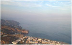 Una vista aérea de un pueblo costero revela edificios entre colinas y el océano bajo un cielo nublado, que recuerda las impresionantes vistas vistas durante un Vuelo en helicóptero sobre el Cañón Tajo de los Vados. - ForTwo - Planes para parejas