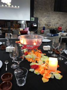 Una refinada escena gastronómica con pétalos naranjas y amarillos, un gran tazón de vidrio, copas de vino y una vela sobre un mantel negro captura la sofisticación de "Descubre la Xocoexperience en el Museu de la Xocolata y podréis convertiros en maestros del chocolate". - ForTwo - Planes para parejas