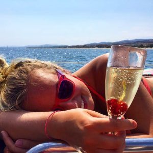 Una mujer disfruta de un "Paseo romántico en velero en Barcelona", sosteniendo una copa de champán con una cereza y luciendo gafas de sol. El idílico paisaje de agua y colinas realza el encanto, capturando la esencia de las tardes soleadas cerca de Barcelona. - ForTwo - Planes para parejas