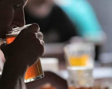 En un ambiente tenue, alguien saborea los ricos sabores de un Taller de cata y degustación de cerveza en Barcelona. Las copas chocan suavemente mientras las figuras borrosas al fondo realzan esta íntima experiencia de cata de cerveza. - ForTwo - Planes para parejas