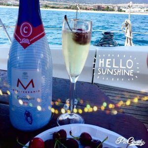 Una botella de vino espumoso y una copa sobre un velero con cerezas, con el mar de Barcelona como fondo, presentan el texto "Hola Sol". Disfruta de esta escena durante tu paseo romántico en velero por Barcelona. - ForTwo - Planes para parejas