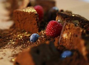 Primer plano de un postre de chocolate con frambuesas, arándanos, cacao y hojuelas de oro sobre madera, una creación que hace eco del capricho de Descubre la Xocoexperience en el Museu de la Xocolata y podréis convertiros en maestros del chocolate. - ForTwo - Planes para parejas