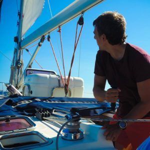 Una persona que navega hábilmente por un paseo romántico en velero en Barcelona, mirando hacia atrás al mar infinito bajo un cielo azul claro, disfruta de un viaje sereno por la costa de Barcelona. - ForTwo - Planes para parejas