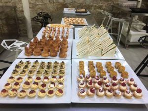 Una variedad de pasteles, sándwiches y mini postres están ingeniosamente dispuestos sobre una mesa en una sala con paredes de piedra, encarnando el placer de "Descubre la Xocoexperience en el Museu de la Xocolata y podréis convertiros en maestros del chocolate". - ForTwo - Planes para parejas