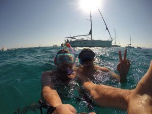 Dos personas con equipo de snorkel sonríen y gesticulan hacia el mar, con barcos y el sol detrás, disfrutando de un "Paseo romántico en velero en Barcelona" a lo largo de la vibrante costa. - ForTwo - Planes para parejas