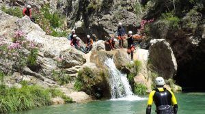 Los participantes, con casco y traje de neopreno, practican barranquismo junto a una pequeña cascada, rodeada de acantilados rocosos y flores rosas, en el impresionante Barranco de Lentegí, cerca de Granada. Disfruta de la experiencia completa con Barranquismo en Granada - Barranco de Lentegí completo. - ForTwo - Planes para parejas