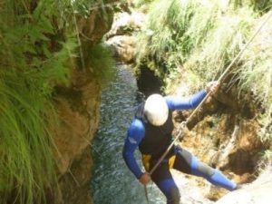 Vestido con un traje de neopreno y un casco, un aventurero desciende con destreza en rápel por el cañón rocoso y herboso del Barranco de Lentegí junto a un arroyo de agua, sumergiéndose en la emoción del "Barranquismo en Granada - Barranco de Lentegí completo" en medio de la pintoresca Granada. - ForTwo - Planes para parejas