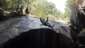 Una persona se desliza por una roca lisa con los brazos en alto, mientras una pareja cercana en el bosque disfruta de una emocionante experiencia con el "Pack multiaventura en pareja en Madrid. - ForTwo - Planes para parejas