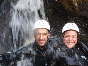 Una pareja con cascos y trajes de neopreno sonríe bajo una cascada, disfrutando de la emoción de su Pack multiaventura en pareja en Madrid. - ForTwo - Planes para parejas