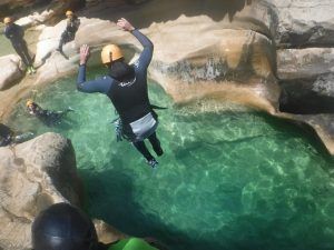 Con casco y traje de neopreno, alguien se sumerge en una piscina de color azul claro entre las rocas en el famoso Pack multiaventura en pareja en Madrid, mientras su pareja y otras personas observan con entusiasmo el emocionante salto. - ForTwo - Planes para parejas