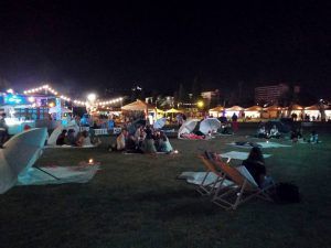 Gente sentada en el césped en un festival nocturno al aire libre, disfrutando de un Menú Montaña Picnic en Barcelona bajo luces de cadena, con carpas y sillas de playa bajo el cielo oscuro. - ForTwo - Planes para parejas