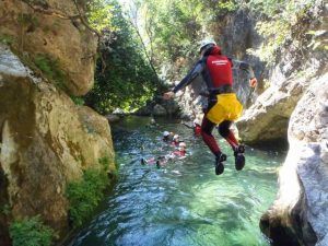 Una persona se lanza a un río cristalino con equipo de seguridad, rodeada de bañistas en un cañón rocoso y boscoso. Esta emocionante aventura de Barranquismo en Granada-Poqueira se desarrolla con el impresionante telón de fondo de la Alpujarra granadina. - ForTwo - Planes para parejas