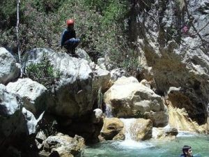 Con un casco rojo, un aventurero se agacha sobre una roca junto a una cascada y una piscina en medio de una exuberante vegetación, todo listo para un emocionante día de Barranquismo en Granada - Barranco de Guardajamas. - ForTwo - Planes para parejas