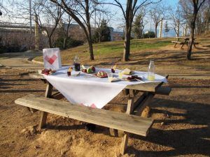 Un picnic soleado en un parque con un mantel blanco, comida variada, bebidas y una bolsa de papel encarna el Menú Mar Salada de Picnic en Barcelona. - ForTwo - Planes para parejas