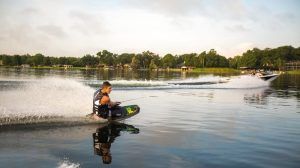 Una persona practica wakeboard con gracia en un lago sereno, utilizando el Kneeboard, una forma diferente de surfear, descubre el Kneeboard en Barcelona, remolcado por una lancha rápida, mientras los entusiastas del kneeboard animan desde la orilla arbolada. - ForTwo - Planes para parejas