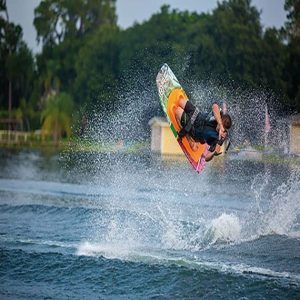 Una persona practica wakeboard con destreza sobre un lago resplandeciente, realizando un impresionante truco aéreo con un chapoteo a cuestas. Altos árboles enmarcan la escena, evocando momentos emocionantes de "Una forma diferente de surfear, descubre el Kneeboard en Barcelona". - ForTwo - Planes para parejas