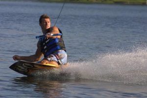 Una persona disfruta de "Una forma diferente de surfear, descubre el Kneeboard en Barcelona" en un lago, rociando agua mientras lleva un chaleco salvavidas azul y sostiene una cuerda de remolque. - ForTwo - Planes para parejas
