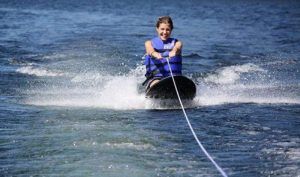 Practicando kneeboard con destreza en un lago, luciendo un chaleco salvavidas azul y creando impresionantes salpicaduras, que recuerdan a "Una forma diferente de surfear, descubre el Kneeboard en Barcelona", con la cuerda tensa marcando el camino. - ForTwo - Planes para parejas