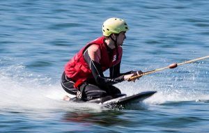 Una persona practica kneeboard en un lago, ataviada con un chaleco salvavidas rojo y un casco verde, agarrando una cuerda de remolque con una euforia similar a la de experimentar "Una forma diferente de surfear, descubre el Kneeboard en Barcelona". - ForTwo - Planes para parejas