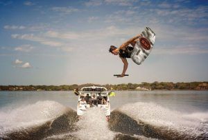 Una persona practica wakeboard en el aire sobre un pintoresco lago, experimentando la emoción de Vive una auténtica experiencia de Wakeboard en Barcelona, con un barco detrás en un día soleado. - ForTwo - Planes para parejas