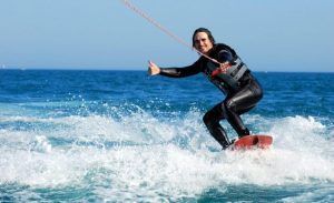 Una persona practica wakeboard en el vibrante océano cerca de Barcelona, luciendo un traje de neopreno y haciendo un gesto de aprobación con el pulgar bajo el cielo soleado: una experiencia verdaderamente inolvidable. Vive una auténtica experiencia de Wakeboard en Barcelona. - ForTwo - Planes para parejas