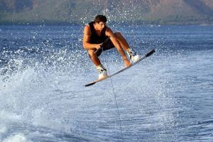 Una persona se desliza por el aire en un emocionante salto de wakeboard con salpicaduras de agua, con un impresionante telón de fondo de montañas y agua azul, una experiencia similar al espíritu aventurero de "Vive una auténtica experiencia de Wakeboard en Barcelona. - ForTwo - Planes para parejas