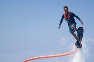 Sobrevolando las cristalinas aguas de Barcelona, una persona que utiliza el "Vuela sobre el agua en Hoverboard o Flyboard en Barcelona" realiza emocionantes acrobacias contra el intenso cielo azul, vistiendo un chaleco salvavidas y pantalones cortos con estampados llamativos. - ForTwo - Planes para parejas
