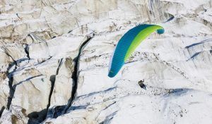 Un parapente con alas azules y verdes se eleva sobre un paisaje montañoso nevado y rocoso, capturando la emoción de "Vuelo en parapente en Granada" en medio de vistas impresionantes. - ForTwo - Planes para parejas